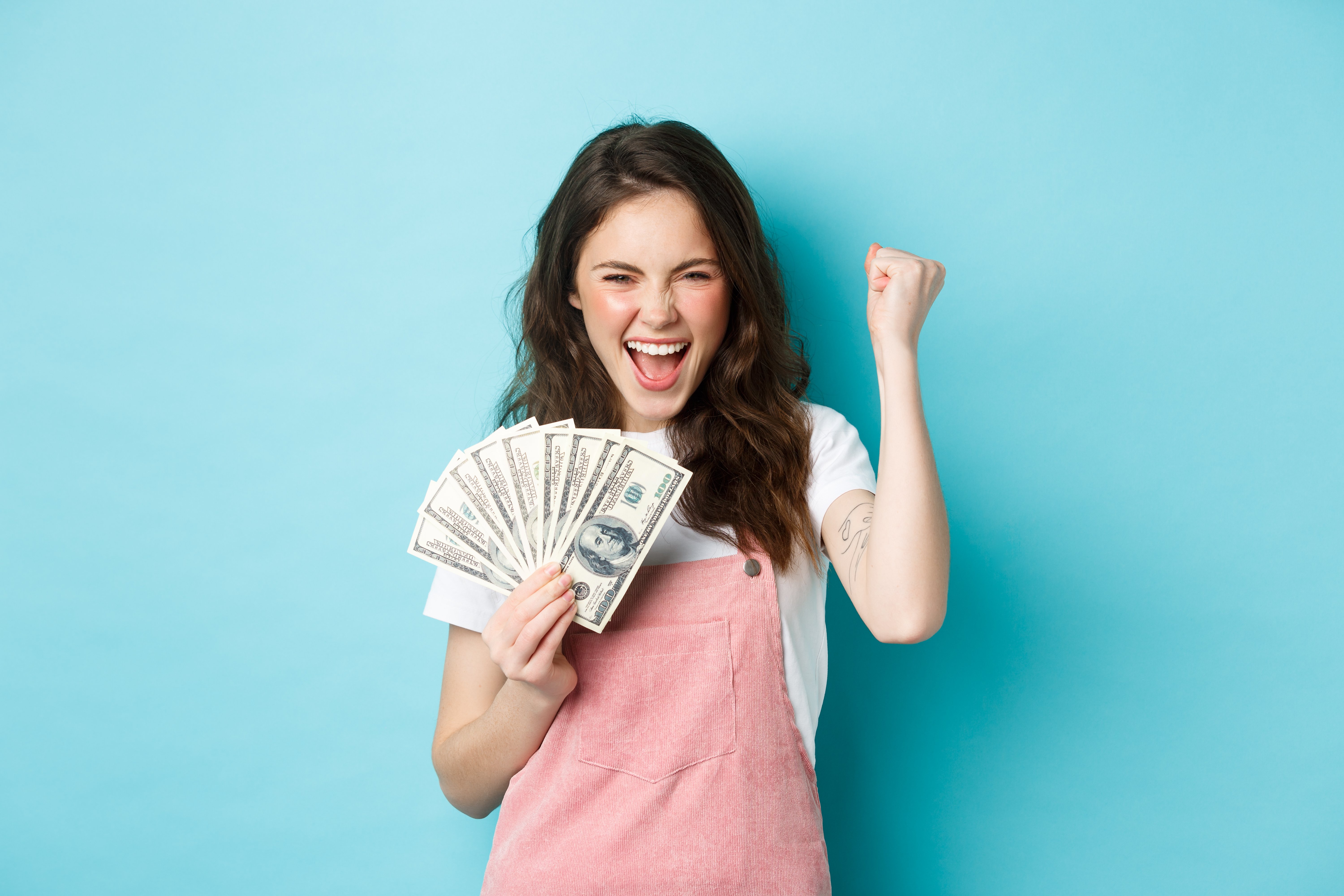 lucky-young-woman-looks-excited-shouting-from-satisfaction-triumph-winning-money-holding-dollar-bills-making-fist-pump-standing-blue-background